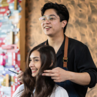 Asian hairstylist male combing and using scissors cut woman's hair. Professional Hairdresser or barber giving treatment and occupation service to smiling young beautiful girl customer in beauty salon.